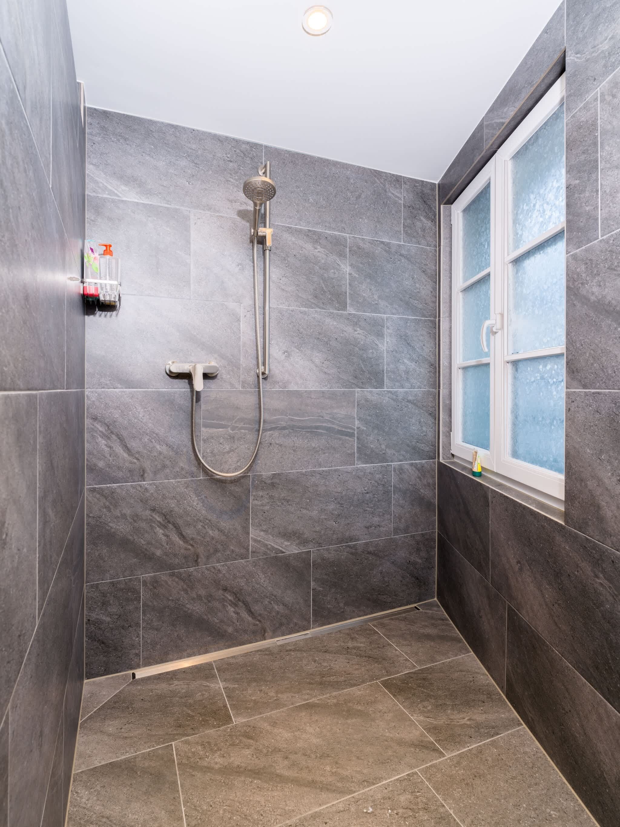 Une douche équipée d’un pommeau de douche à main et d’un robinet mural, une fenêtre avec du verre dépoli et une petite étagère contenant des articles de toilette.