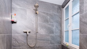 A shower with a handheld showerhead and a wall-mounted faucet, a window with frosted glass, and a small shelf with toiletries.