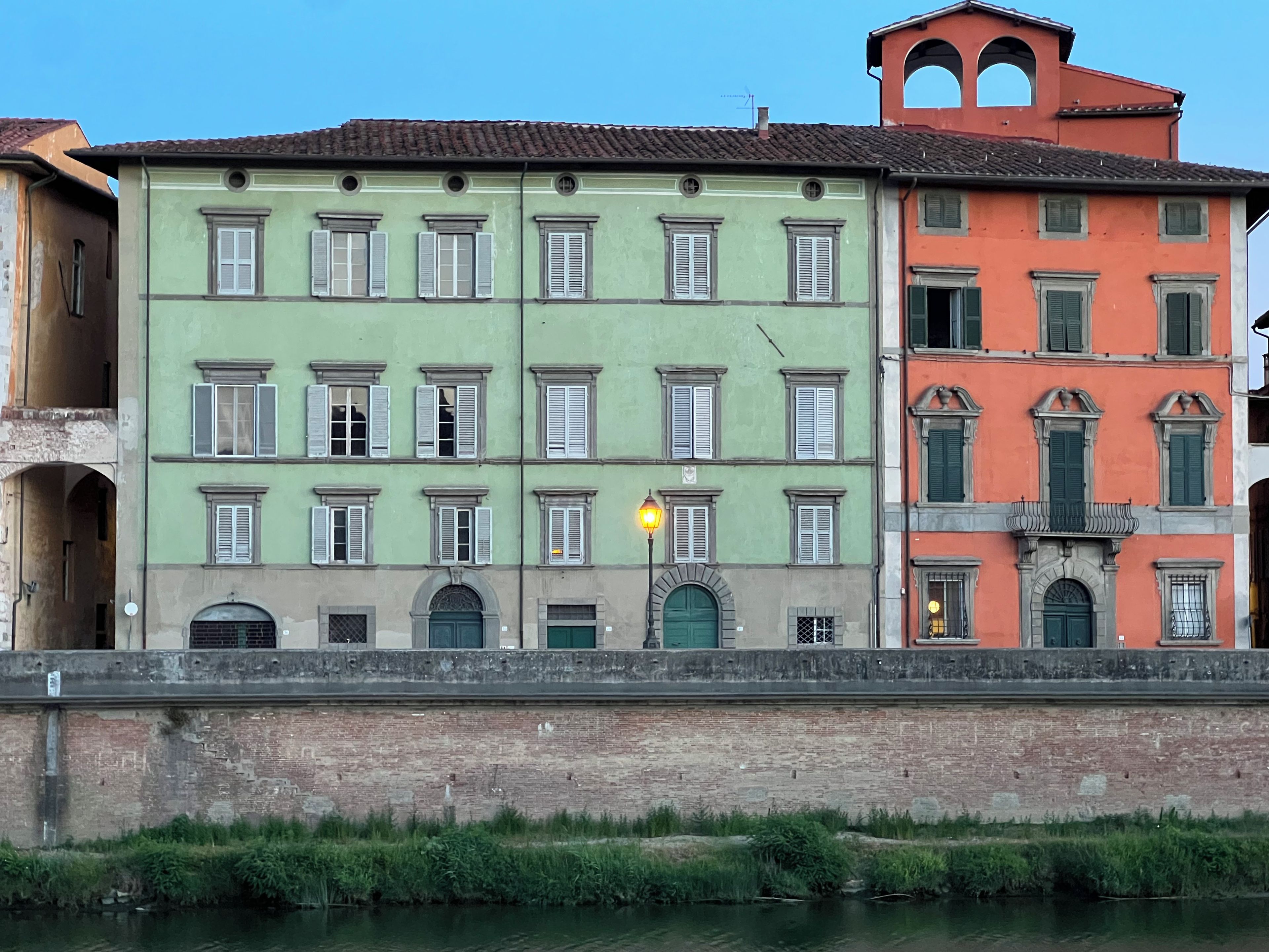 A row of historic buildings with multiple windows and doors, a brick wall in the foreground, and a canal with greenery in the background.