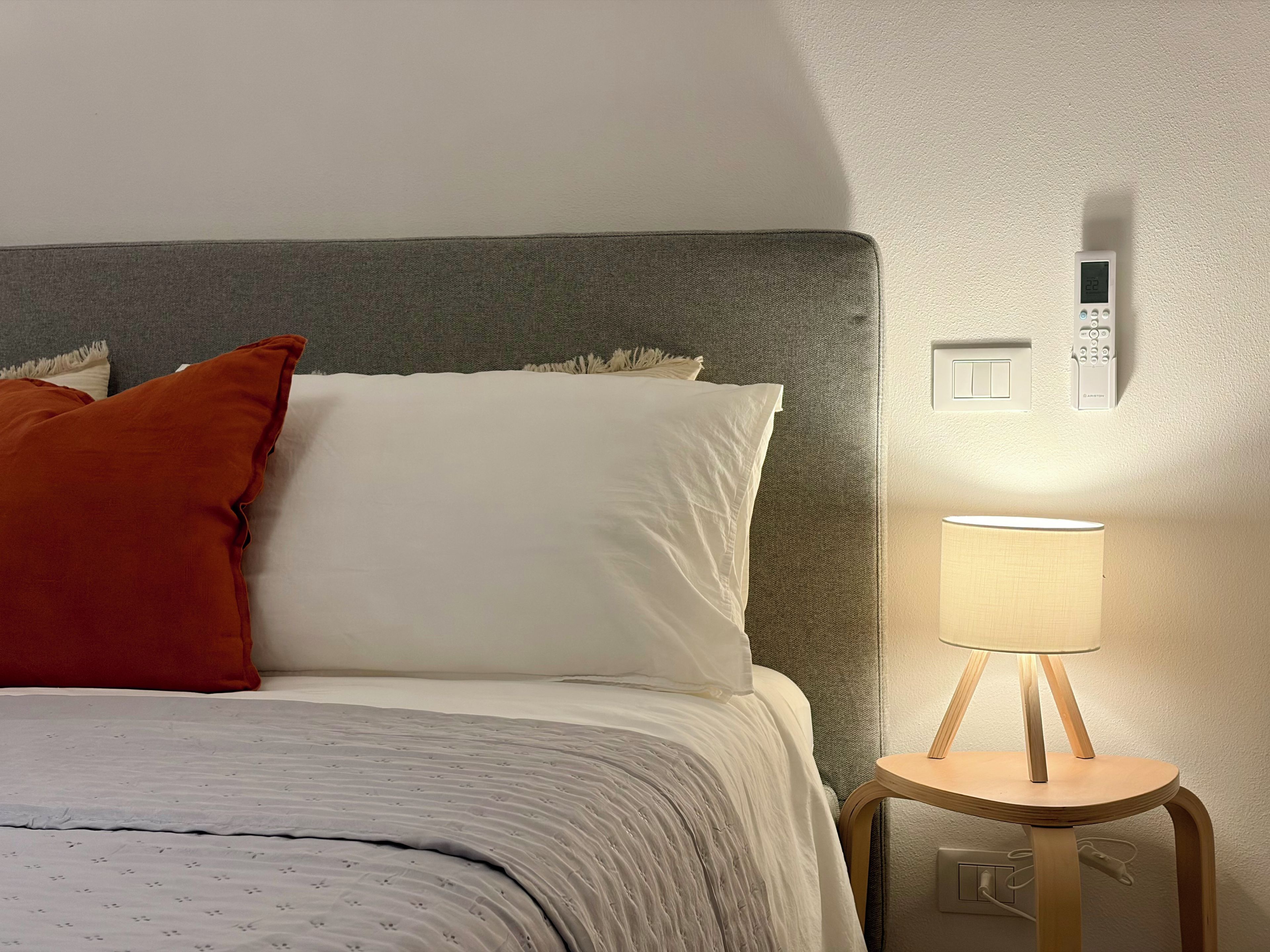 A bed with white and orange pillows, a bedside lamp, and a thermostat on the wall.