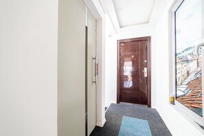 A hallway with a wooden door, a white wall, and a window with a cityscape view.
