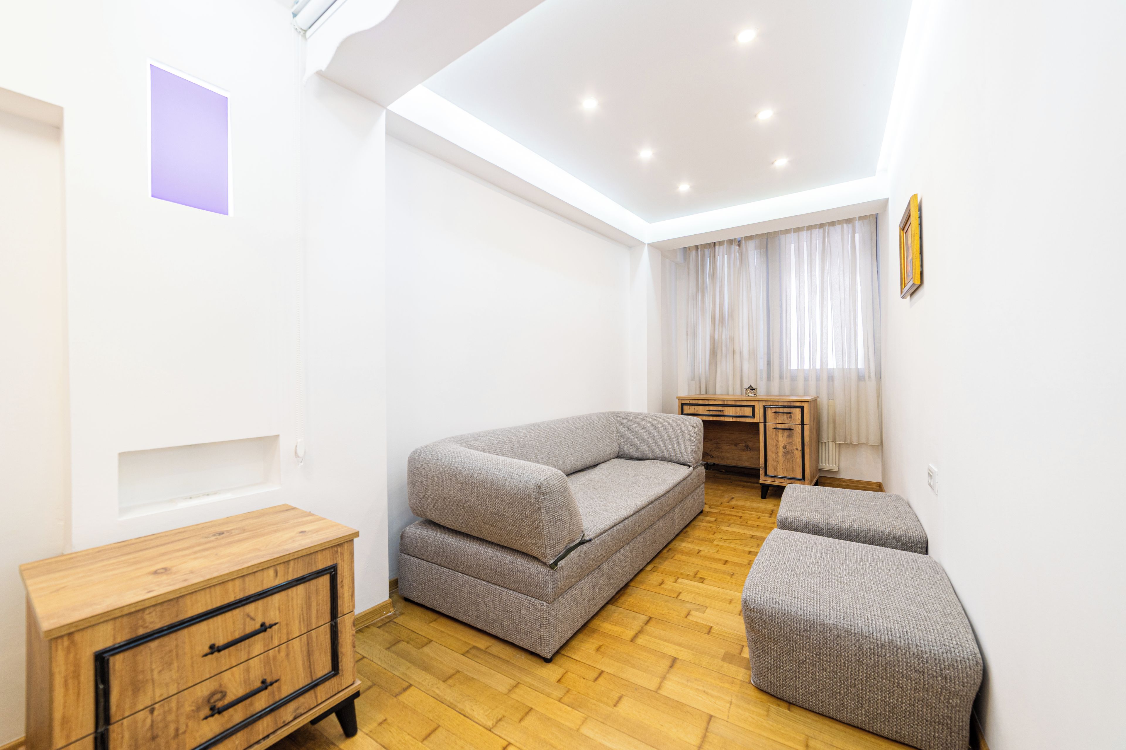 Uma sala de estar moderna com um sofá, um banquinho, um armário de madeira e uma escrivaninha com uma cadeira.