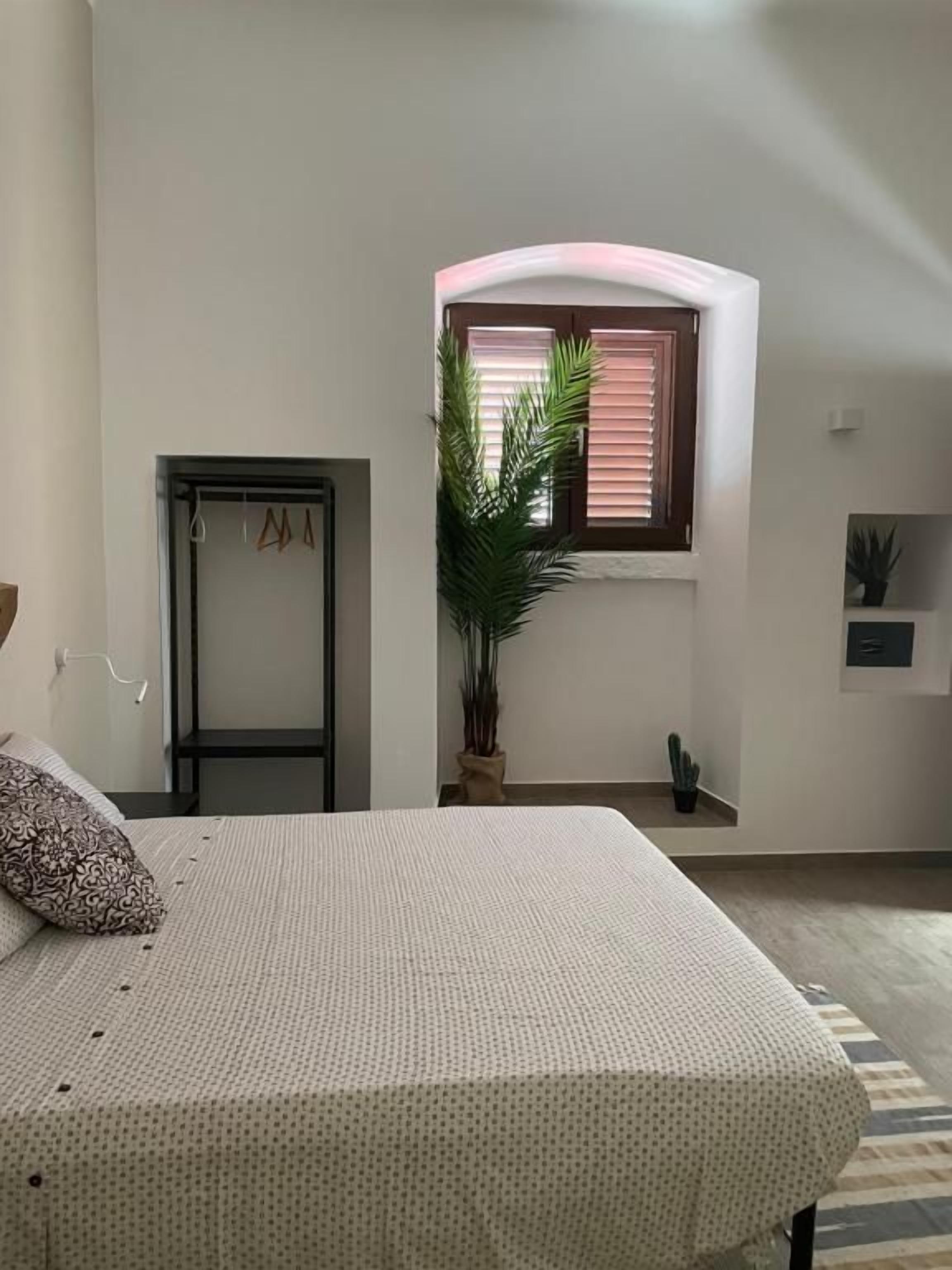 Una camera da letto con un letto, una finestra con persiane, una pianta in vaso, una porta e un piccolo ripiano.