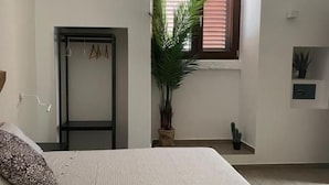 Ein Schlafzimmer mit einem Bett, einem Fenster mit Jalousien, einem Topfpflanzen, einer Tür und einem kleinen Regal.