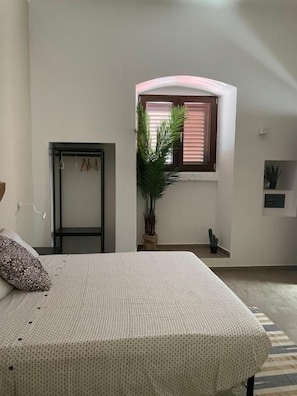 A bedroom with a bed, a window with blinds, a potted plant, a door, and a small shelf. - L angolo delle bugie (TURI)
