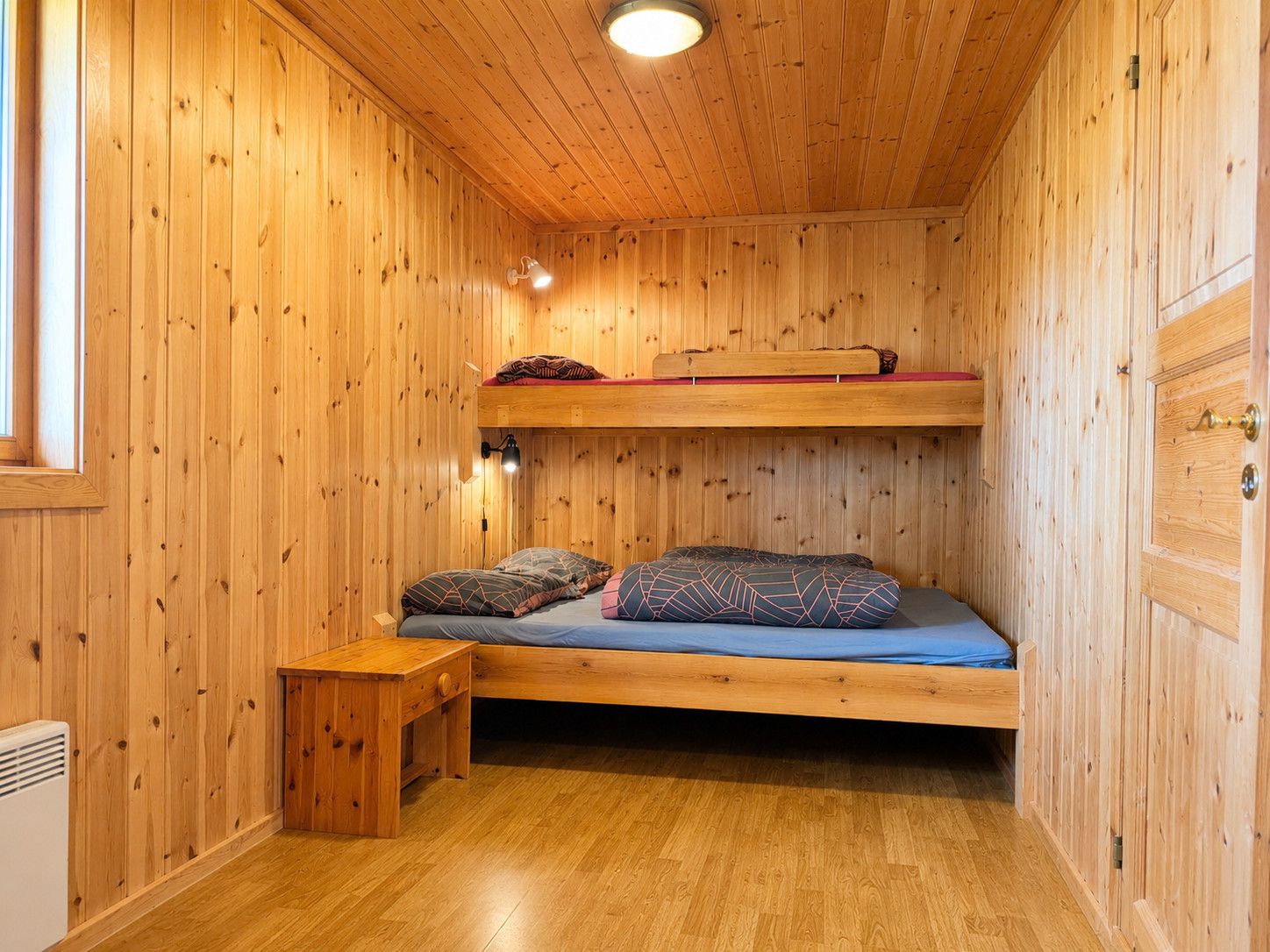 Habitació de fusta amb un llit superposat, una taula de fusta petita i una làmpada fixada a la paret.