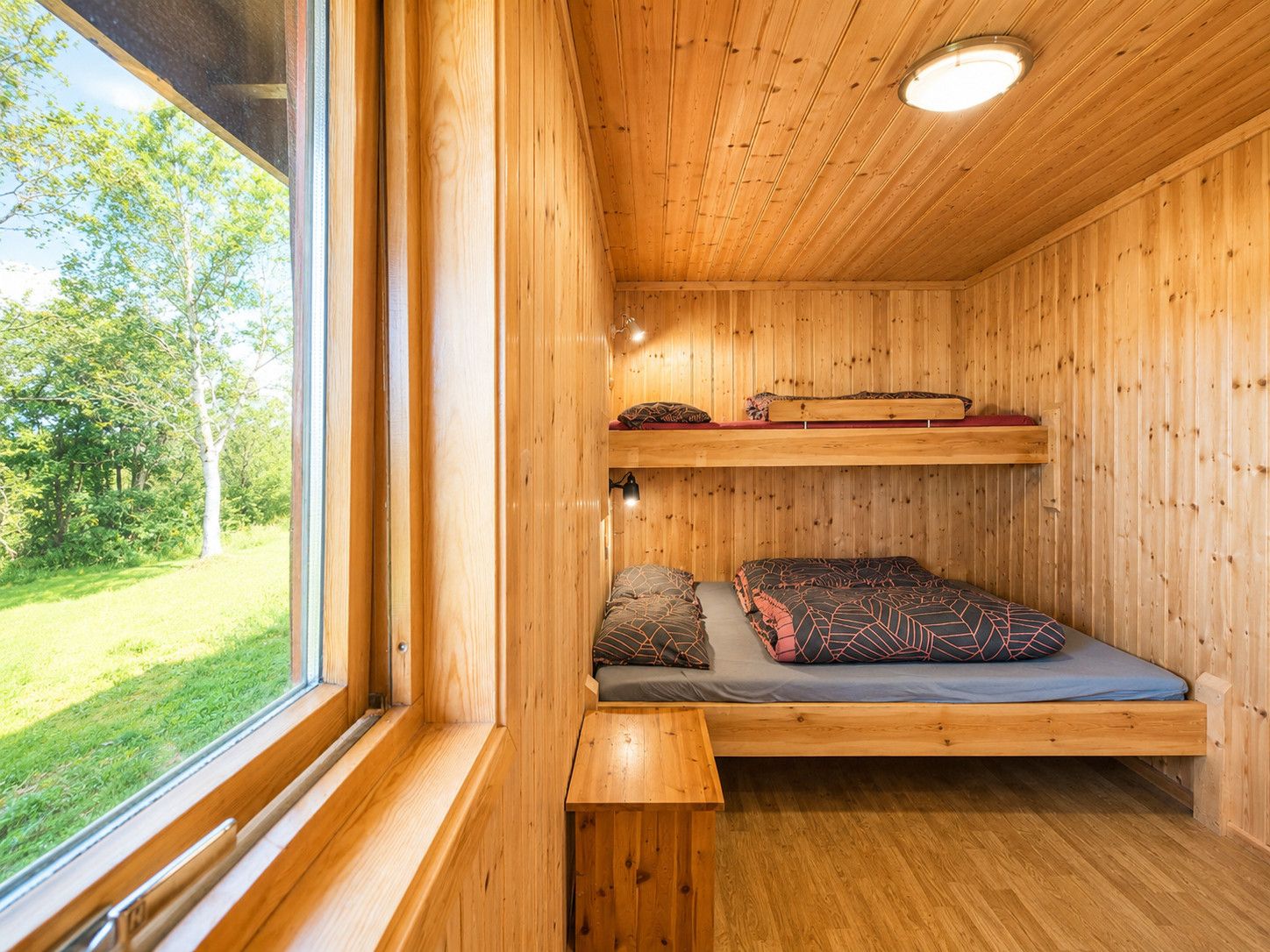 L’intérieur d’une cabane en bois, avec un lit superposé, un banc et une fenêtre donnant sur une zone herbeuse.