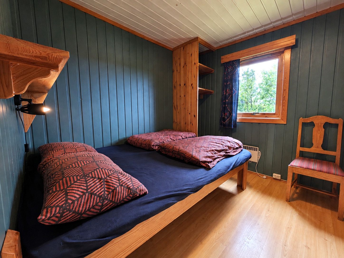 Uma cama de solteiro com almofadas estampadas, uma cadeira de madeira e uma janela com vista para o verde.
