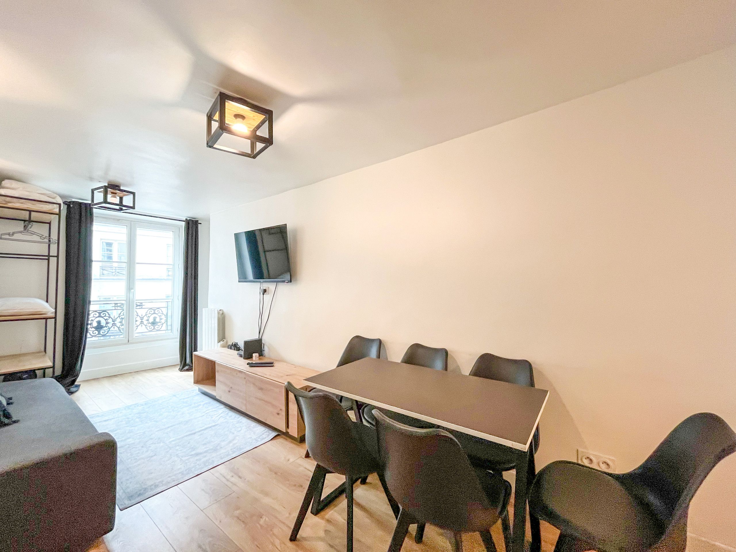 A room with a dining table, chairs, a TV mounted on the wall, and a window with curtains.
