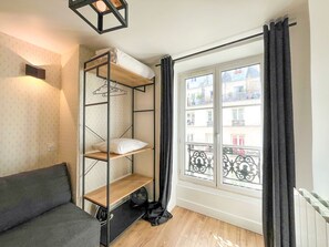 Ein kleines Zimmer mit einem Etagenbett, einer Schlafcouch, einem Heizkörper und einem Fenster mit Blick auf Gebäude.