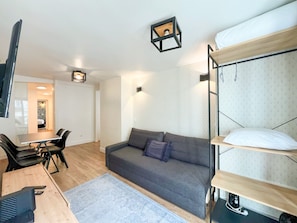 Ein kompakter Wohnbereich mit einer Couch, einem Esstisch mit Stühlen, einem Schlafstockbett mit Matratze und einem kleinen Teppich.