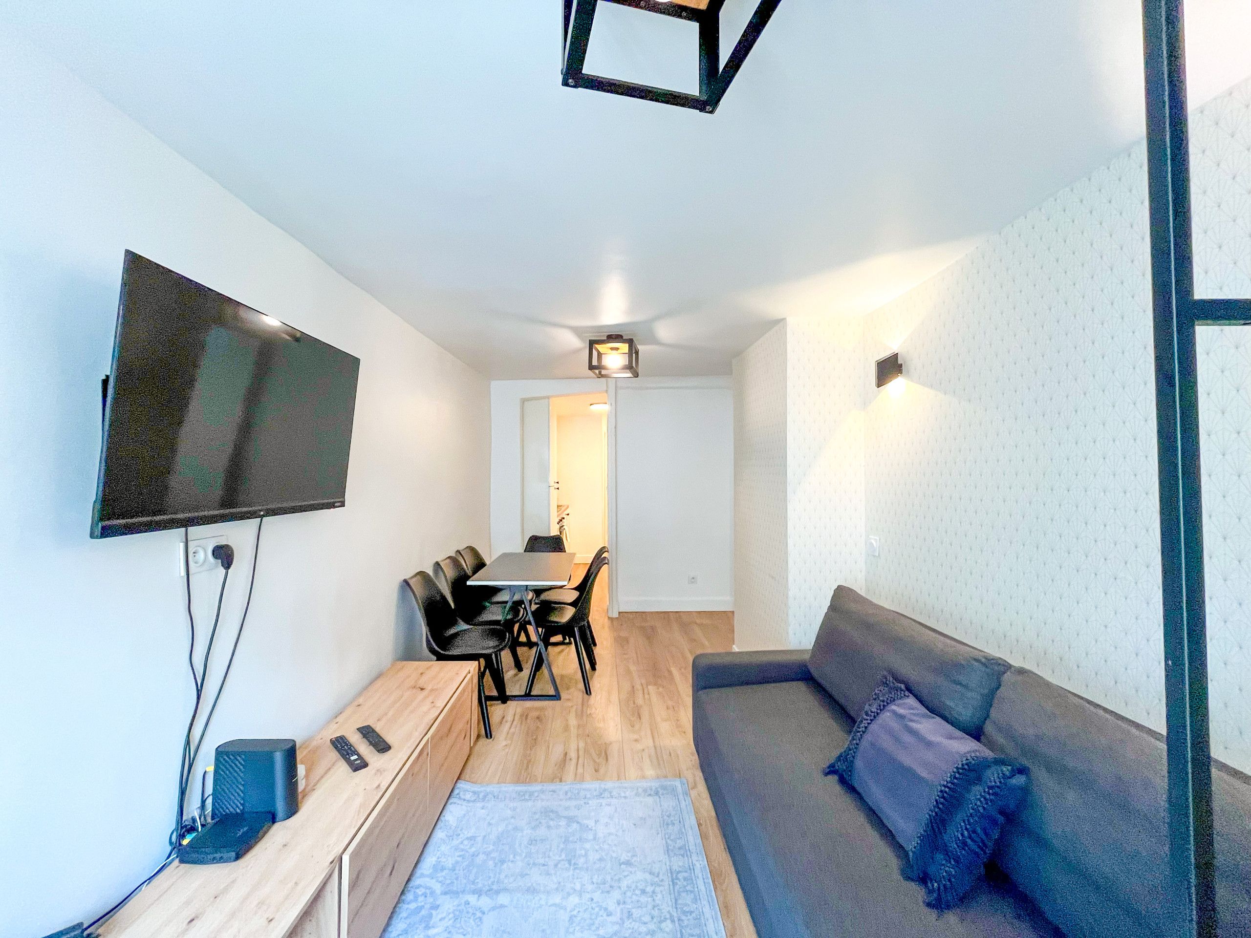 Un espace de vie moderne comprenant un téléviseur à écran plat fixé au mur, un canapé, une table à manger avec des chaises et une petite table d’appoint en bois.