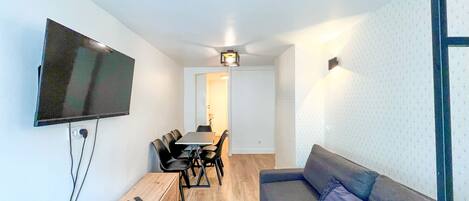 Un espace de vie moderne comprenant un téléviseur à écran plat fixé au mur, un canapé, une table à manger avec des chaises et une petite table d’appoint en bois.