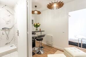 Et moderne badeværelse med bruser, toilet og håndvask. Der er en kommode med et spejl og en vase med blomster. Værelset har trægulv og hvide vægge.