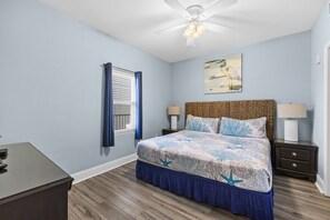 Une chambre à coucher avec un lit, un ventilateur de plafond, une fenêtre avec des rideaux, une table de chevet, une lampe et un tableau au mur.