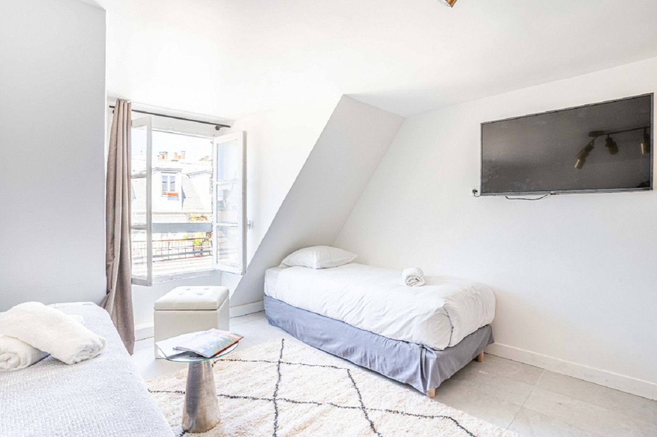Une chambre à coucher avec un lit, une petite table, une fenêtre avec des rideaux et un téléviseur fixé au mur.