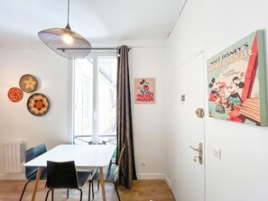 A dining area with a table and chairs, a hanging light fixture, a window with curtains, and a Mickey Mouse poster on the wall. - 146 - Urban Cosy Flat in Paris (París)