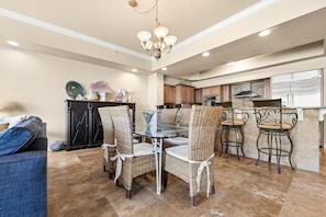A spacious living area with a dining table surrounded by wicker chairs, a dark wood cabinet, and a kitchen in the background. - Spacious Coastal Retreat | Complimentary Beach Service (Destin)