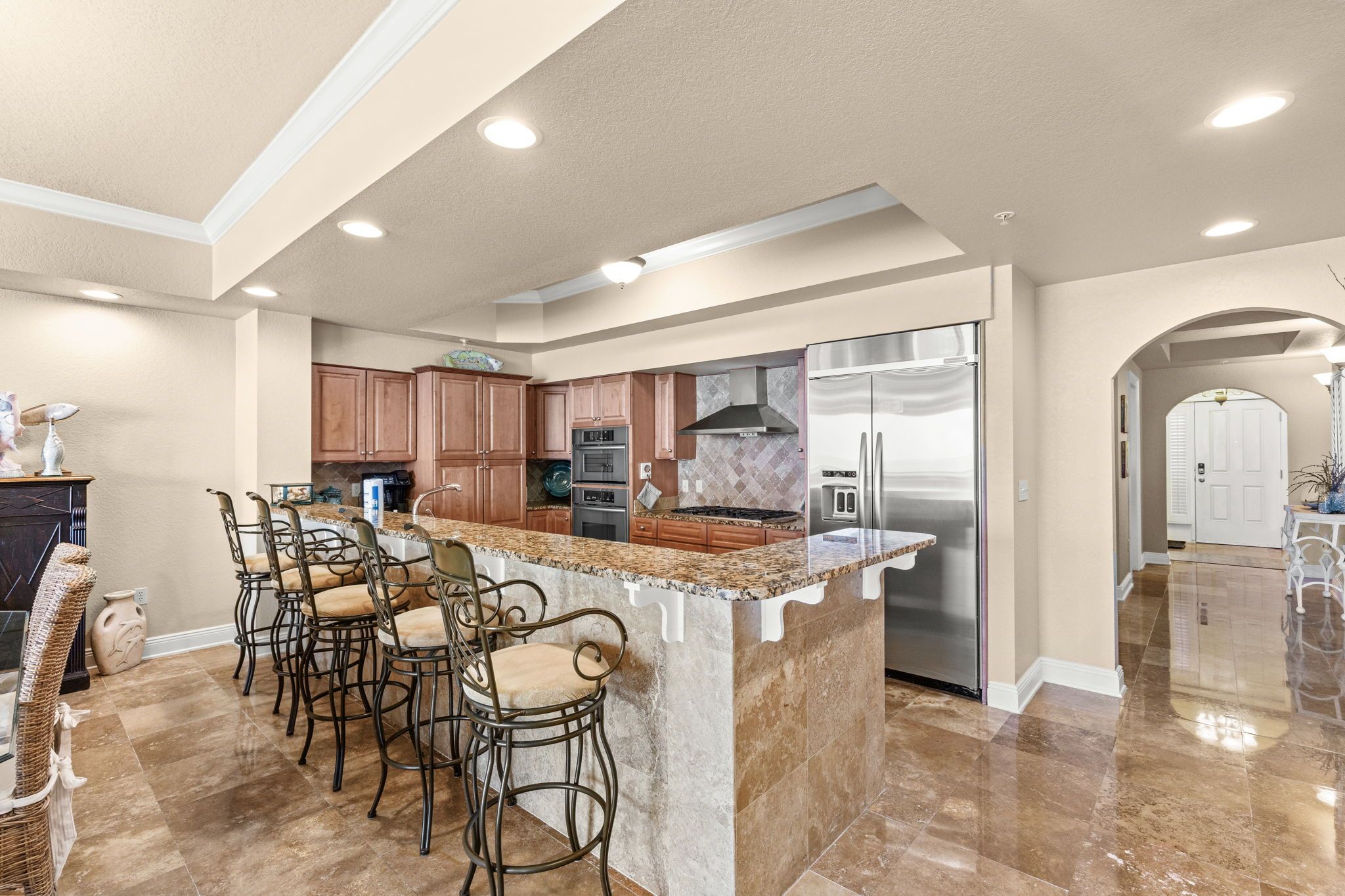 A modern kitchen with a central island, bar stools, stainless steel appliances, and a polished tile floor.