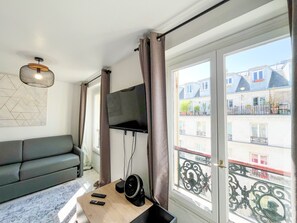 A living room with a sofa, a flat-screen TV mounted on the wall, a wooden coffee table with a speaker, and a large window with a view of buildings. - 143 - Urban Charming Flat in Center of Paris (París)