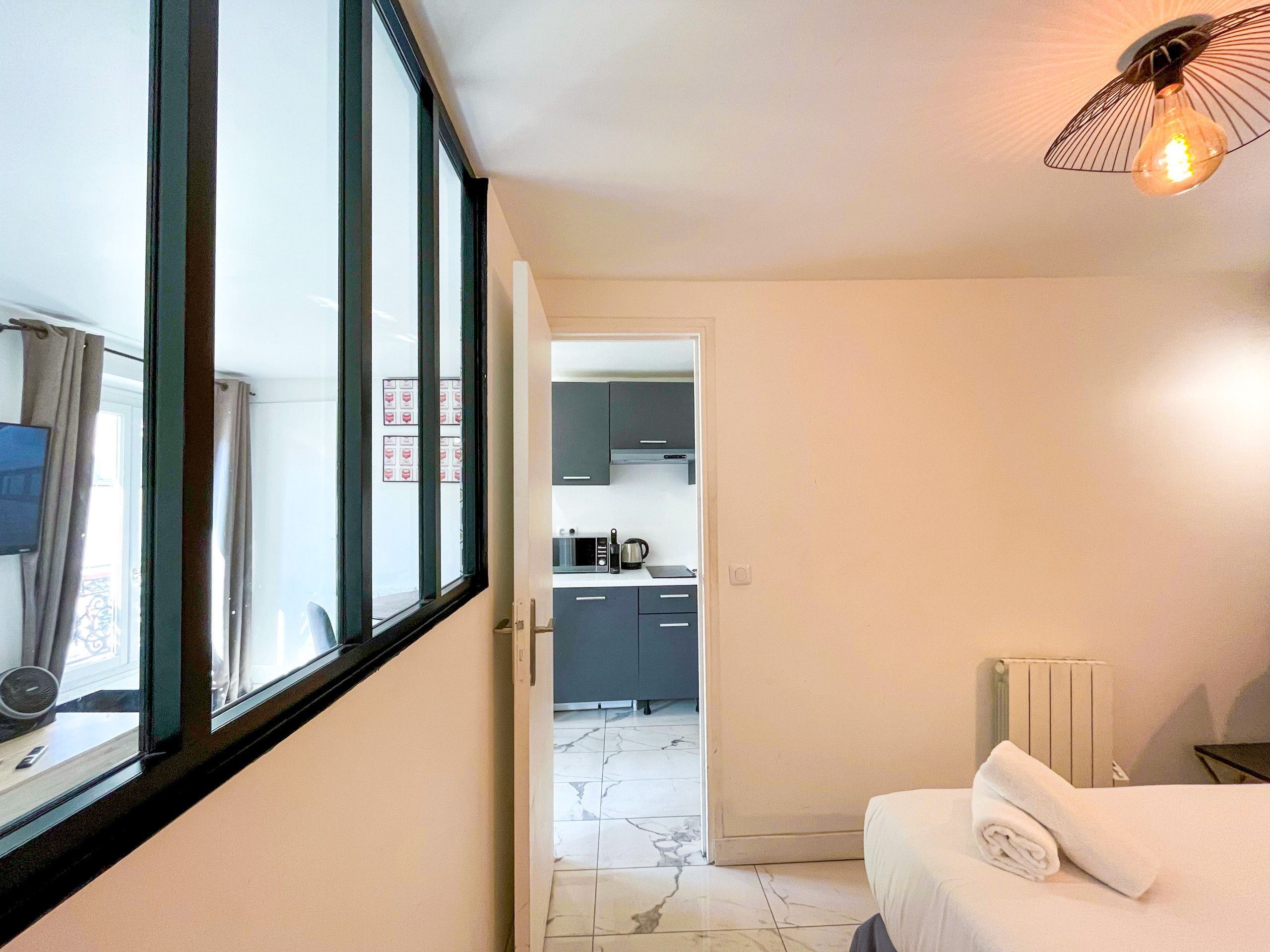A hotel room with a bed, a window with curtains, a kitchen area visible through an open door, and a unique light fixture.
