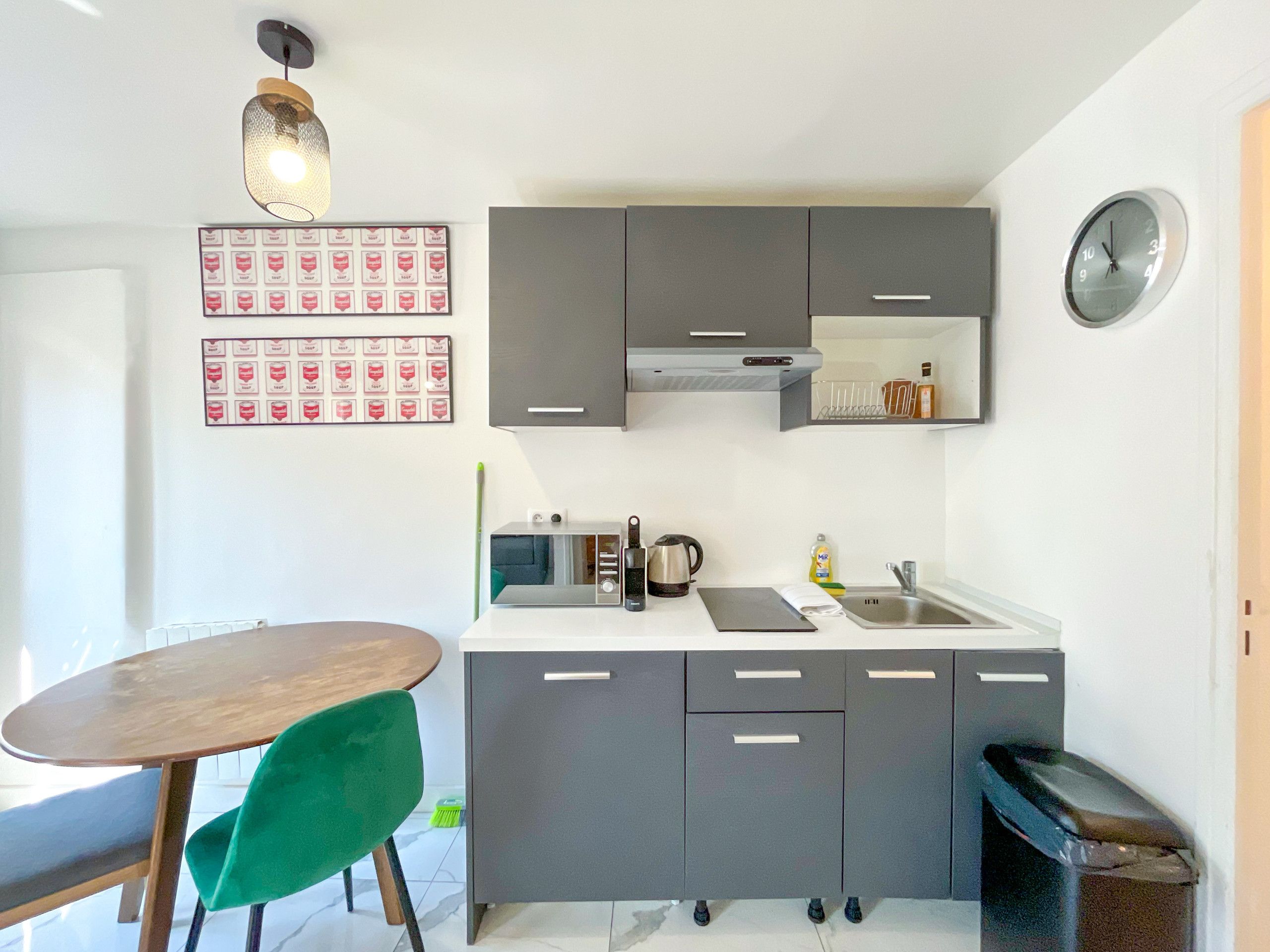 A compact kitchen with grey cabinets, a white countertop, a green chair, and a wall-mounted clock.