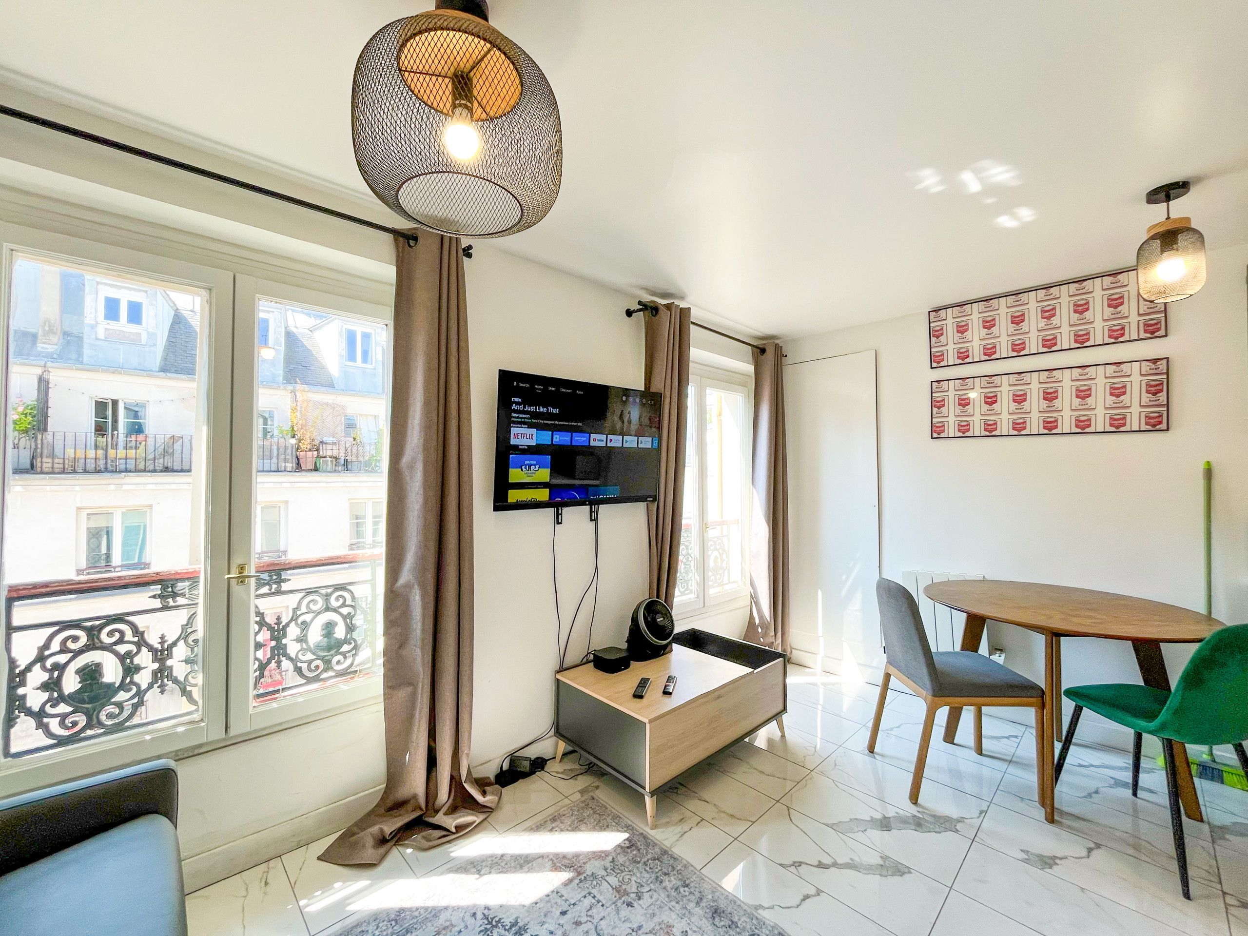 A room with a TV mounted on the wall, a wooden table with chairs, a window with curtains, and a balcony view.