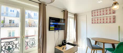 A room with a TV mounted on the wall, a wooden table with chairs, a window with curtains, and a balcony view.