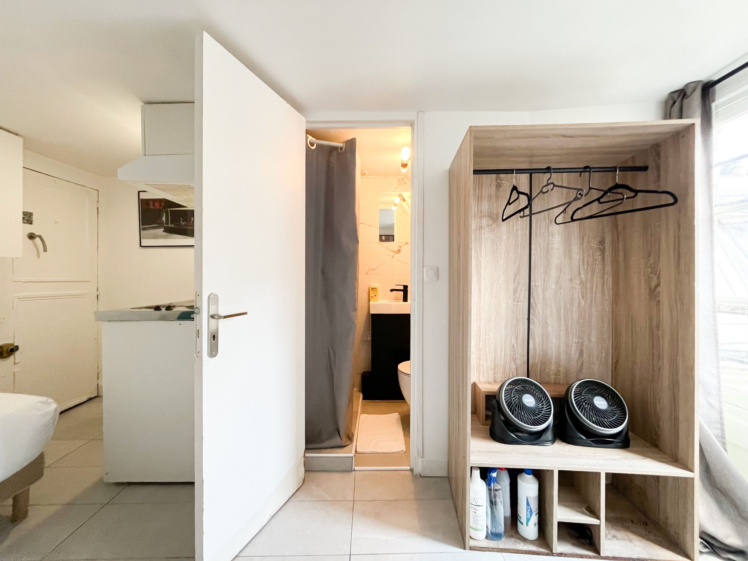 Une chambre d’hôtel compacte comprenant un lit, une petite kitchenette, une salle de bain avec des toilettes et un lavabo, ainsi qu’une armoire en bois équipée d’un ventilateur et de cintres.