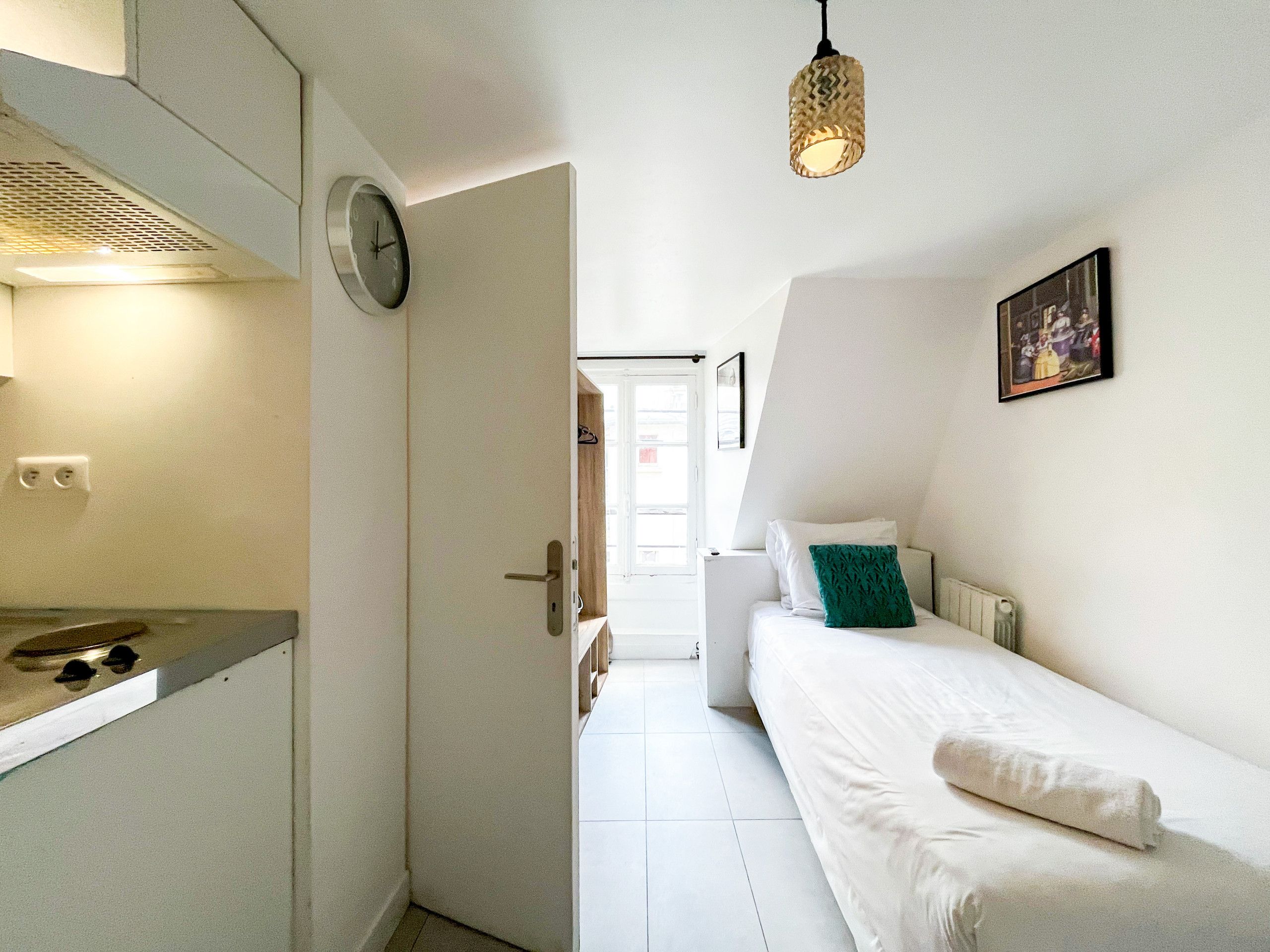 A compact room with a kitchenette, a bed with a green pillow, and a painting on the wall.