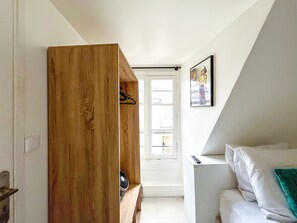 Ein Zimmer mit einem Holzschrank, einem weißen Bett mit einem türkisfarbenen Kopfkissen, einem Fenster mit Blick und einem gerahmten Bild an der Wand.