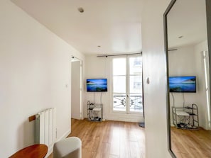 Ein Zimmer mit Holzboden, einem weißen Heizkörper, einem Sessel, einem an der Wand befestigten Fernseher und einem Fenster mit Blick auf Gebäude.