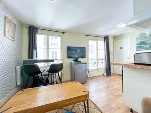 A compact living space with a kitchenette, a dining area with a table and chairs, a television, and a large window with curtains. - 163 - Urban Studio in Paris (París)