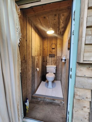 A small, rustic wooden bathroom with a toilet, a hanging light fixture, and a towel rack. - Badgers Den at The Secret Garden (Ashford)
