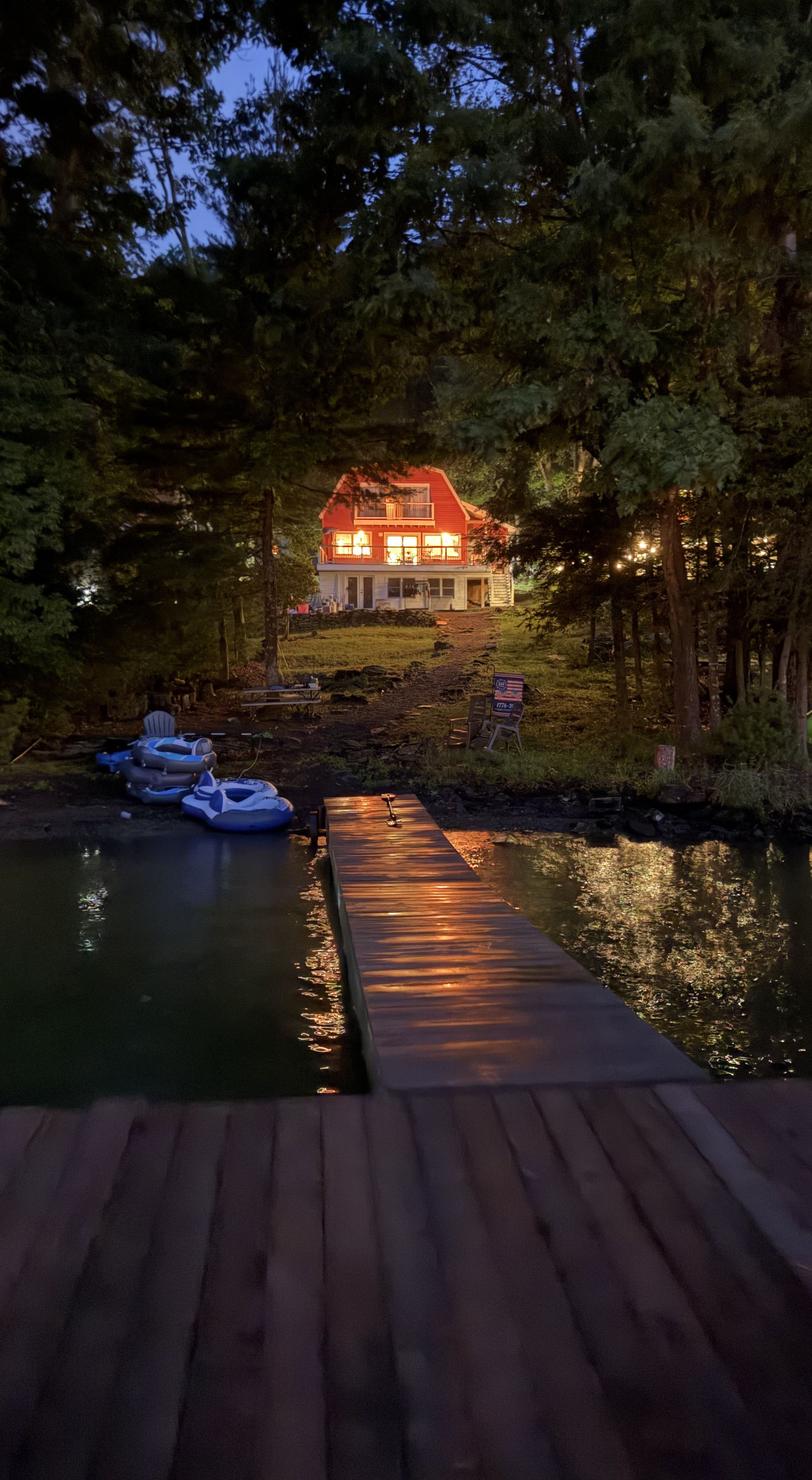A wooden dock leading to a house with a red roof, surrounded by trees and a calm body of water.