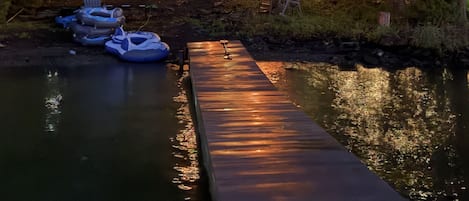 Une jetée en bois mène à une maison avec un toit rouge, entourée d’arbres et bordée par une étendue d’eau calme.