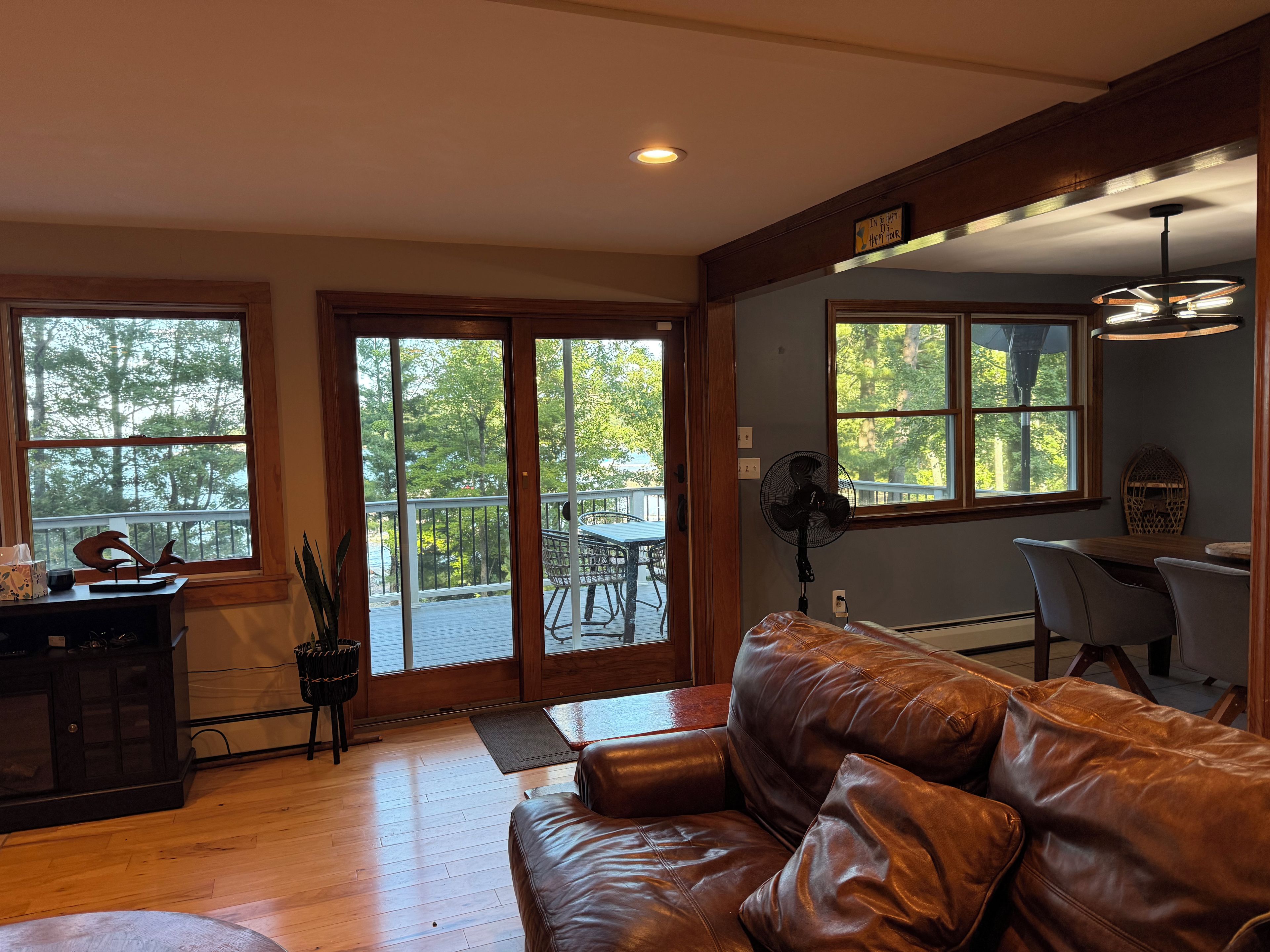 A spacious living room with a leather couch, a dining area with a table and chairs, a fan, and large windows offering a view of trees.