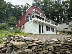 A two-story house with a red roof and white siding, a balcony, and a stone wall in the foreground. - Waterfront house on Lake Wallenpaupack w/ private dock, sleeps 9, dogs allowed! (Tafton)