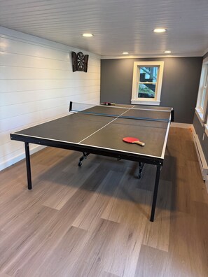 Une table de ping-pong avec un filet, une raquette et une balle, dans une pièce dont le sol est en bois et qui possède une fenêtre.