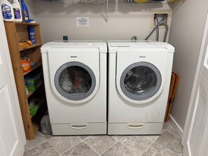 Deux machines à laver Bosch blanches se trouvent dans une buanderie, avec une étagère remplie de produits de nettoyage.