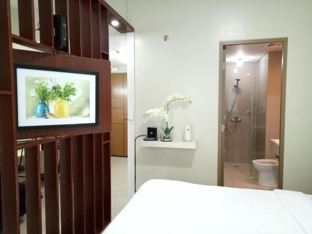 Ett hotellrum med en säng, en tv som är monterad på en vägg av trä, en liten hylla med dekorativa föremål och en duschhörna.