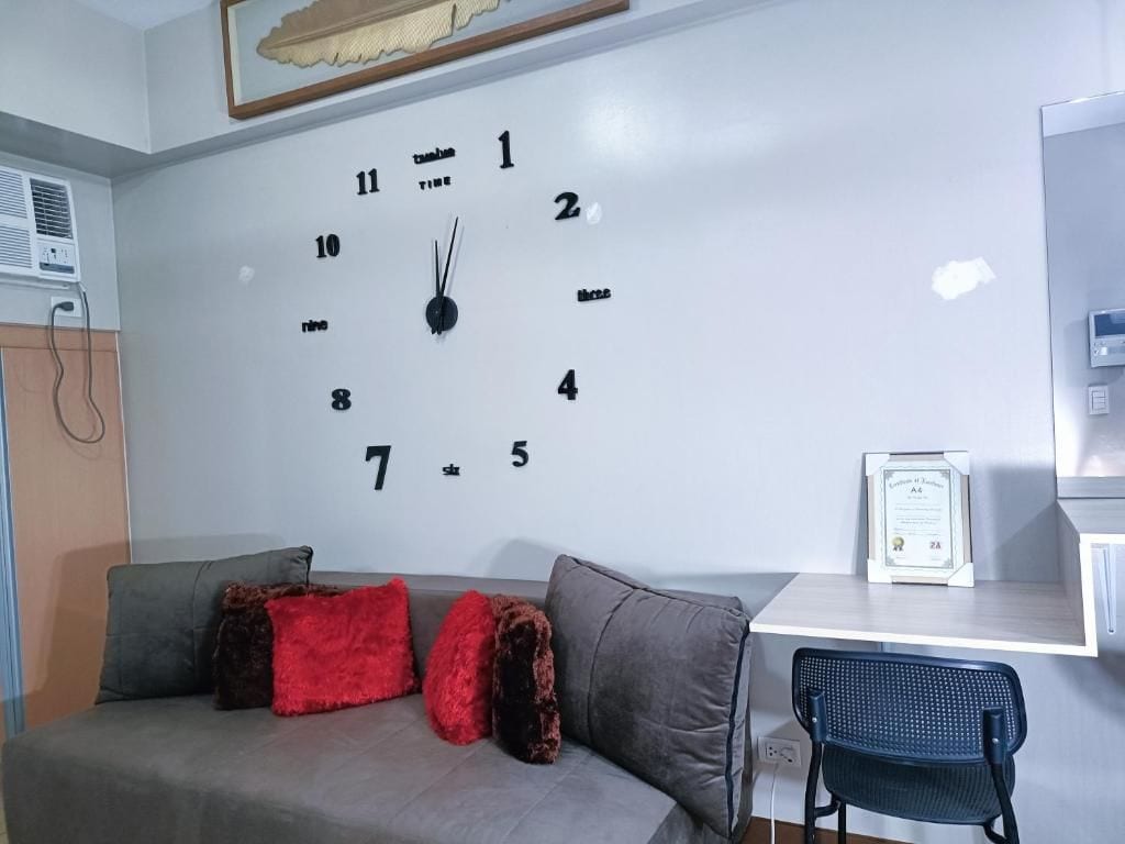 A room with a white wall clock, a grey sofa with red and brown cushions, a small table with a framed certificate, and a black chair.