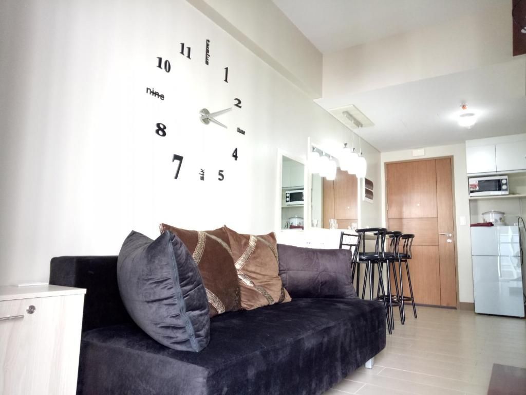 A living room with a black sofa, a wall clock, and a kitchen area in the background.