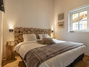 Ein Schlafzimmer mit einem Kopfende im Blockholzmuster, einem Bett mit weißer Bettwäsche und einer gemusterten Tagesdecke, einem Nachttisch mit Lampe und einem Fenster mit Blick auf Gebäude.