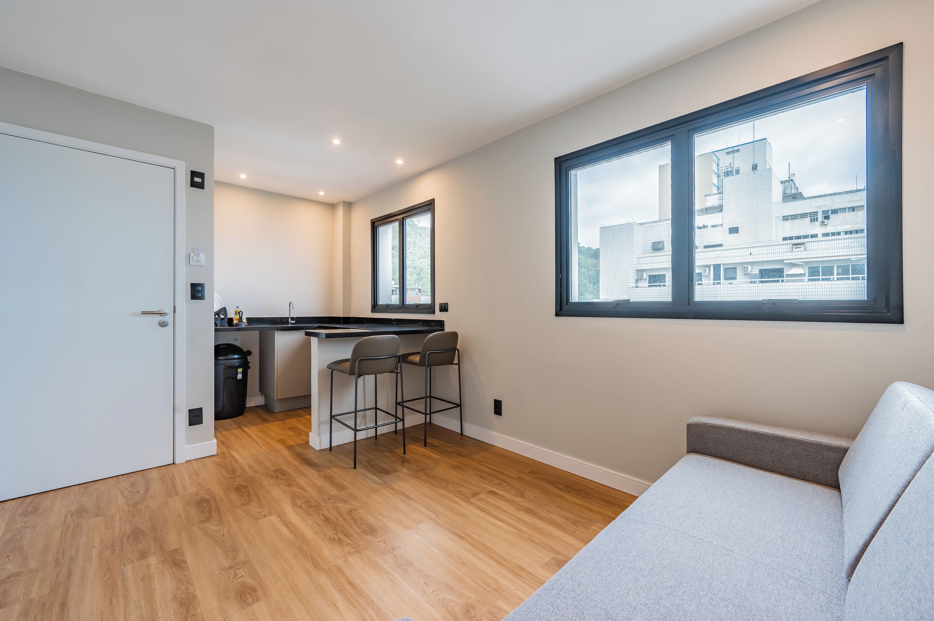 A modern apartment with a kitchenette, a dining area with bar stools, a large window with a city view, and a sofa.