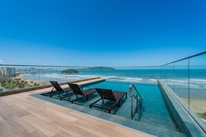 Une piscine à débordement située sur le toit, avec des chaises longues, offrant une vue sur une plage et l’océan.
