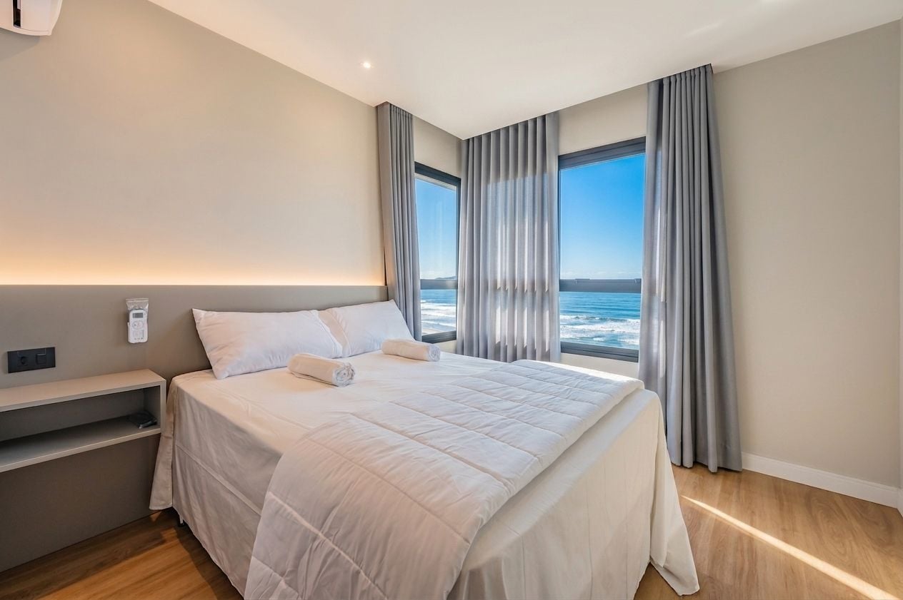 Una habitación de hotel moderna con una cama grande, una mesita de noche y vistas al mar a través de amplias ventanas.