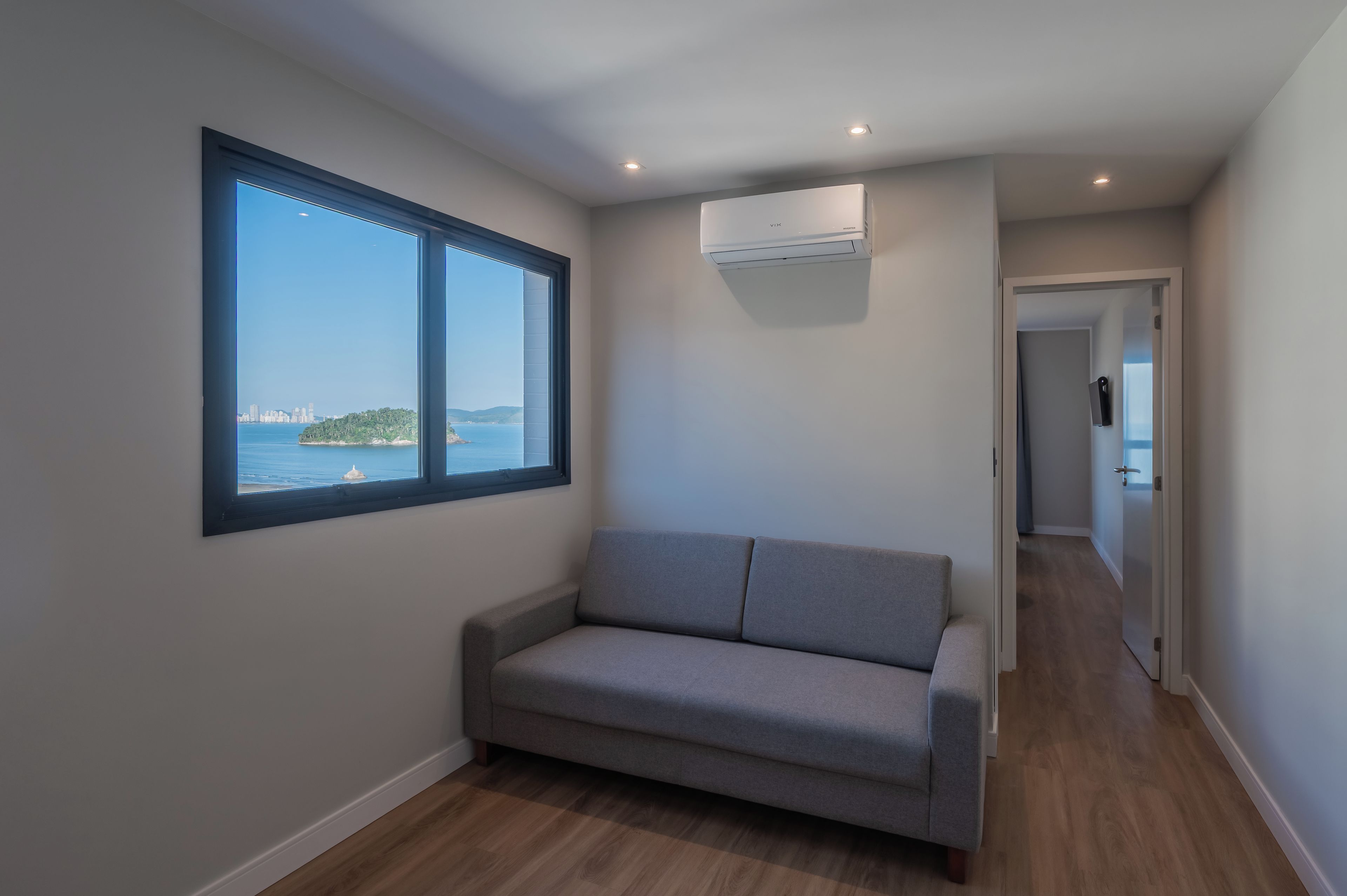 Een kamer met een grijze bank, een groot raam met uitzicht op een stad en water, en een airconditioning aan de muur bevestigd.