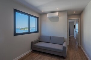 Una habitación con un sofá gris, un ventanal con vistas a la ciudad y al agua, y un equipo de aire acondicionado en la pared.