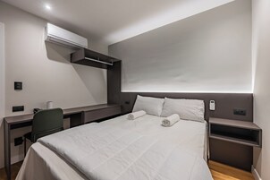 Quarto de hotel moderno com cama, mesas de cabeceira, escrivaninha e aparelho de ar condicionado.